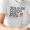 “Solitude Couch Quilt Book #mypriorities” typography graphic with a reading character illustration printed on a white t-shirt | Smolt
